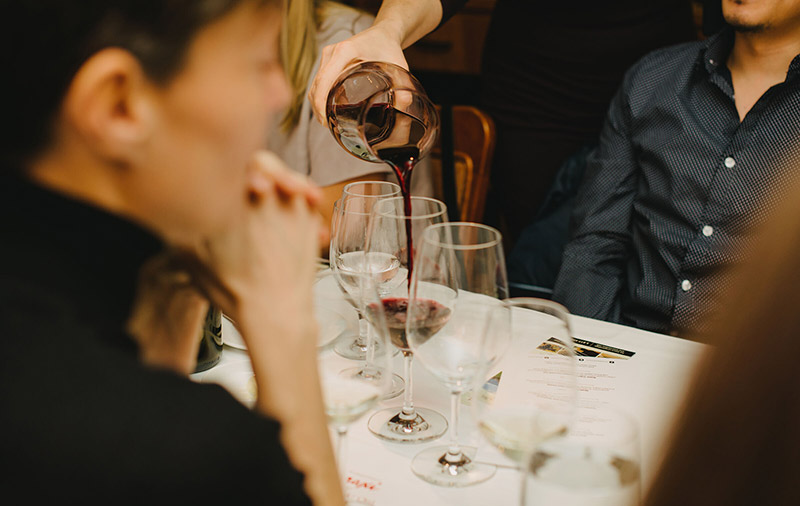 A person's hand with chopsticks picking up food from a dish on a white linen-covered table laden with glasses at Cornucopia Whistler